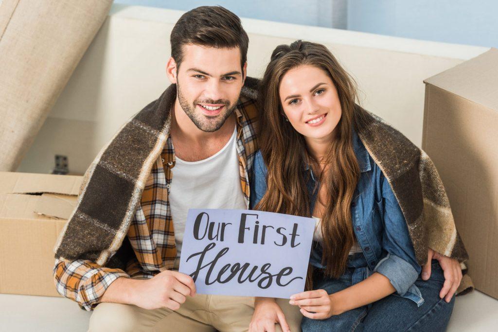 First-time home buyers holding &ldquo;Our First House&rdquo; sign in new home surrounded by moving boxes