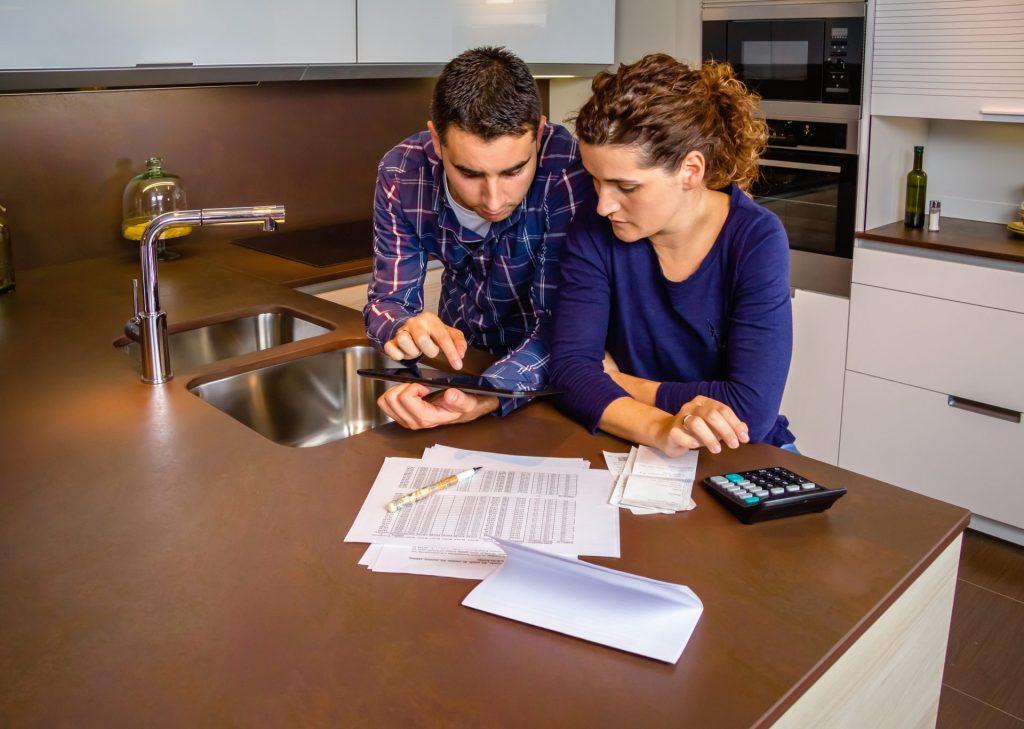 Couple reviewing household finances and mobile banking options together at a kitchen counter using a tablet and calculator